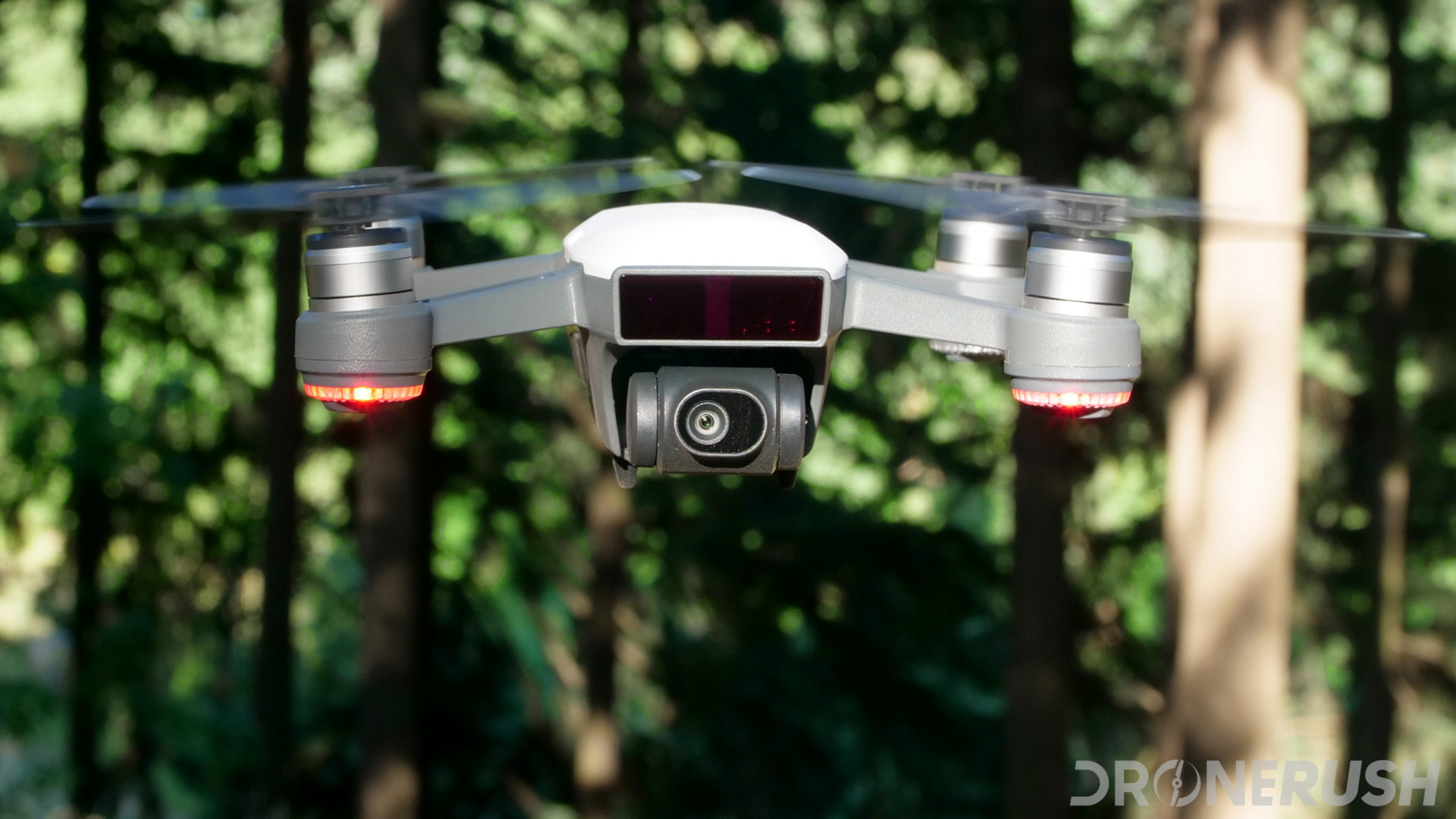 Image of a white DJI Spark hovering in front of trees, the drone is facing the camera so to show off the drone's camera, this is our DJI Spark camera review.